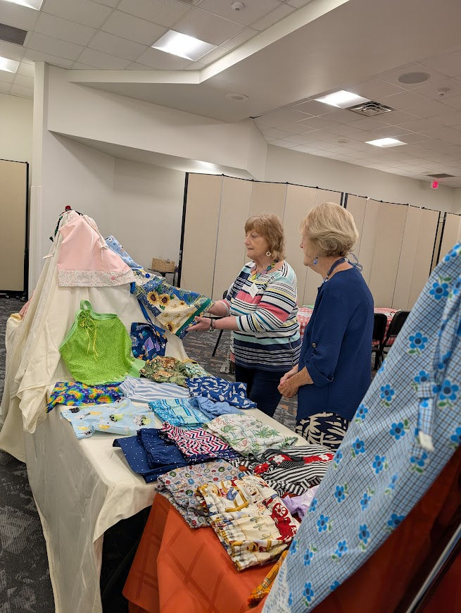 Two older women browse colorful handmade cloths on a table in a conference room with beige dividers.