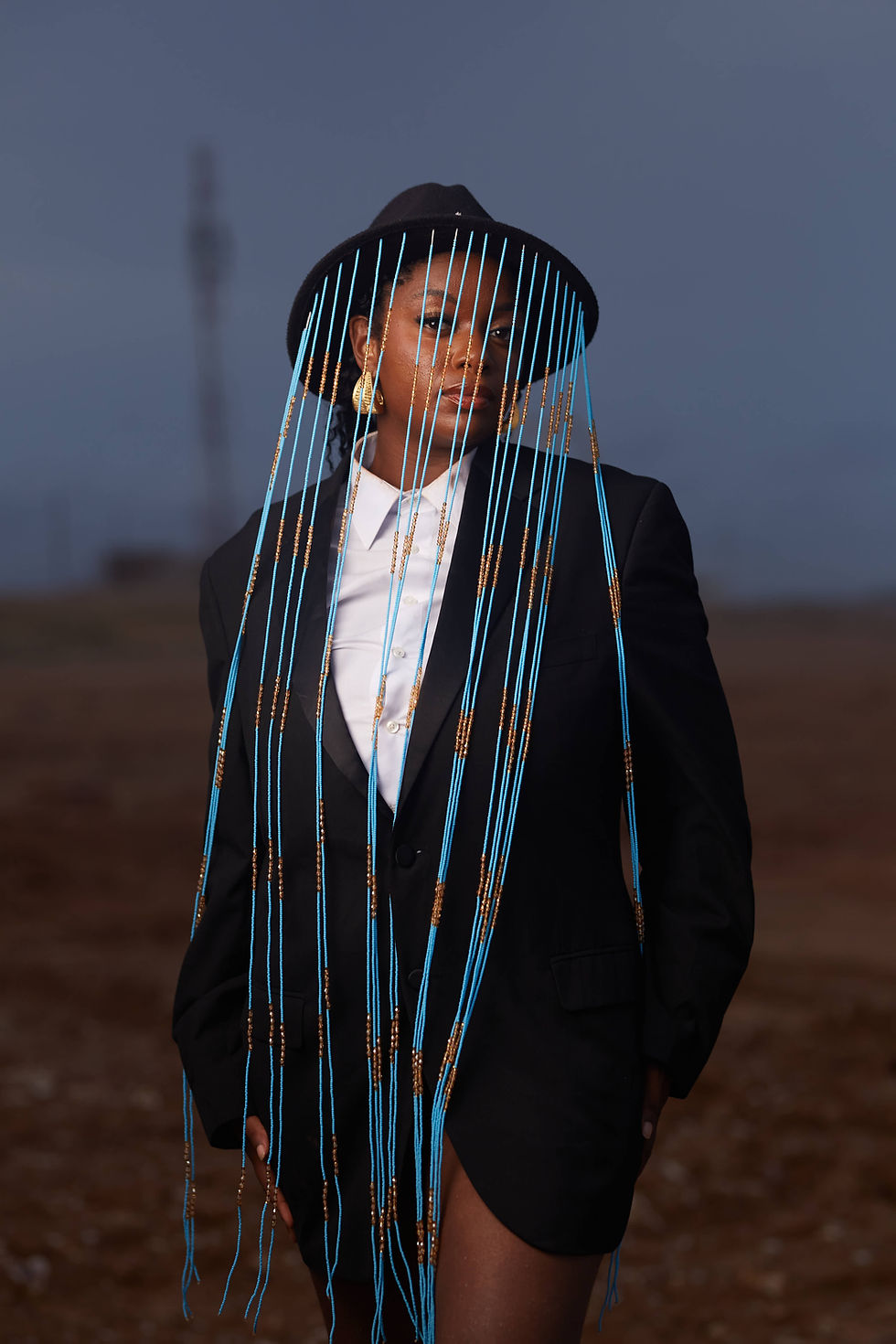 Zakisha in a black suit and wide-brim hat adorned with long blue beaded fringe, standing against a dramatic outdoor backgroun