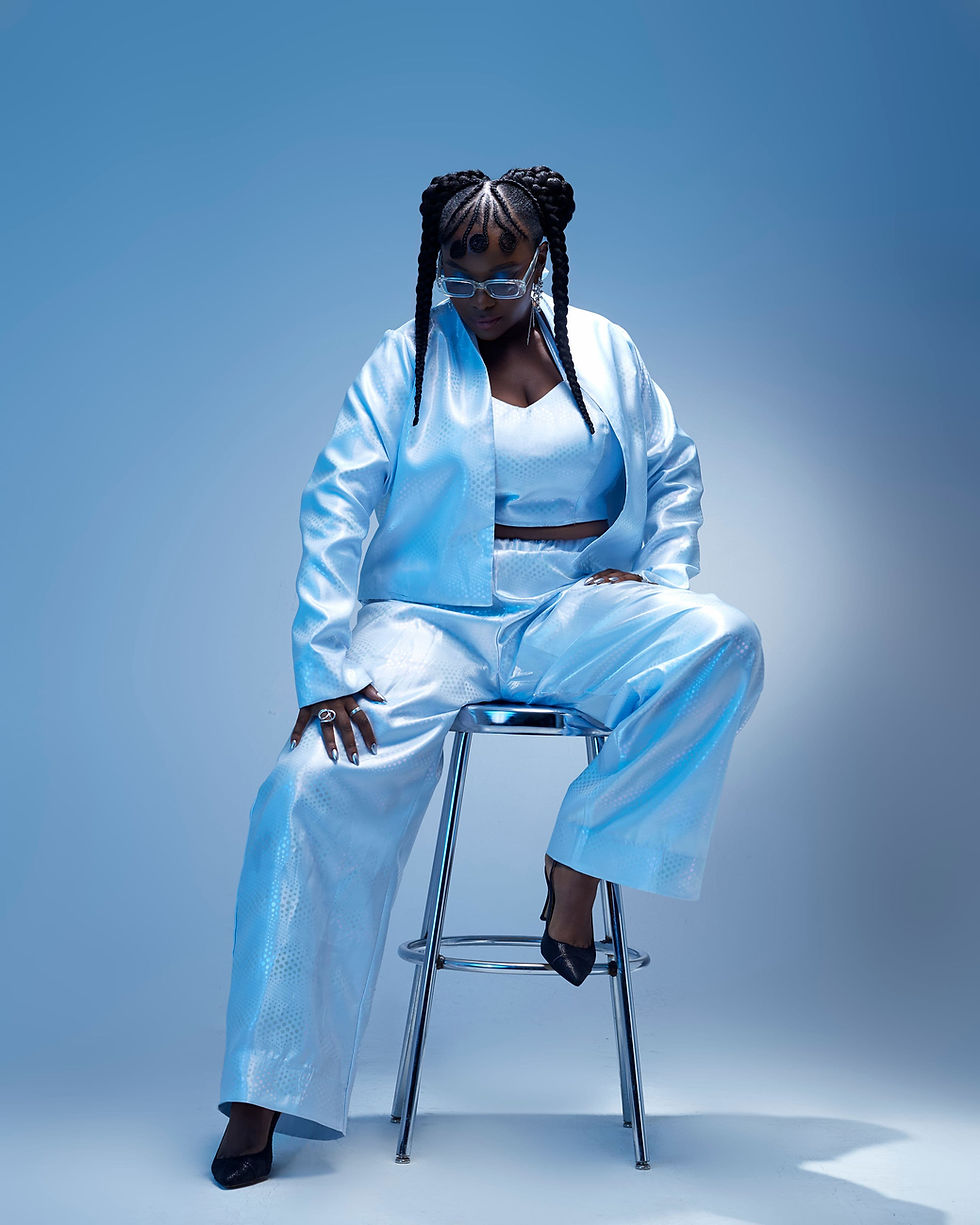 Zakisha Brown seated confidently on a chrome stool, dressed in a reflective light blue outfit with statement braids and clear