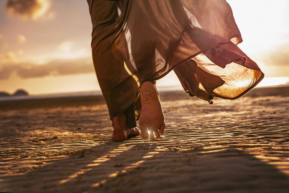 Eine Person in einem fließenden Kleid geht barfuß am Strand entlang, beleuchtet von der warmen Abendsonne