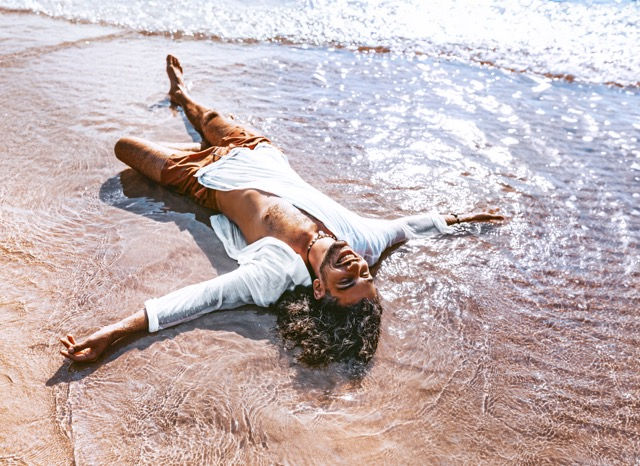 Person liegt entspannt im flachen Wasser am Strand, Arme und Beine ausgestreckt, sonniges Wetter