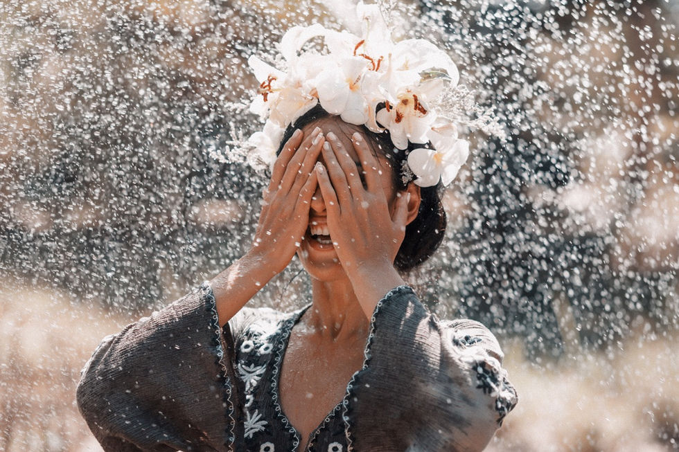 Frau mit Blumenkranz, hält Hände vor das Gesicht, Wassertropfen überall, fröhliche Stimmung, Naturhintergrund.