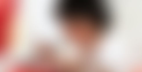 Foto de Geoneide Brandão pintando, segurando uma paleta coberta de tintas predominantemente vermelhas e amarelas. Nota-se o pincel mergulhando na tinta e o uso intenso de vermelhos e ocres.