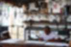 Diogum, um homem negro de camisa branca, assina, concentrado, serigrafias dispostas sobre uma mesa. O ambiente é um ateliê, com várias obras de arte feitas de ferro penduradas e espalhadas ao redor. Há diversos objetos e ferramentas de trabalho visíveis, sugerindo um espaço criativo e artesanal.