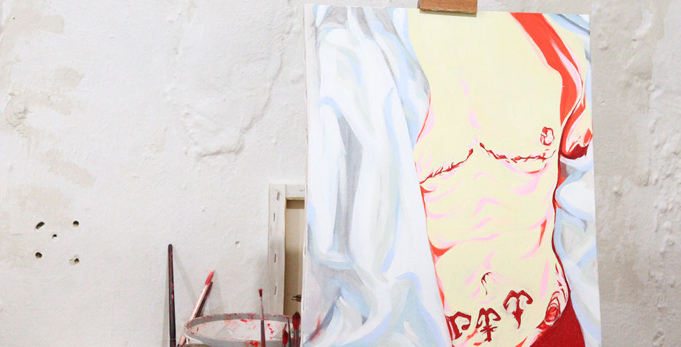 A imagem mostra um cavalete com uma tela em processo de pintura, apoiada contra uma parede branca texturizada. A pintura retrata o torso nu de uma pessoa por um tecido azul e branco. As cores predominantes são tons claros, com destaque para vermelho, rosa e amarelo pálido. Há tatuagens visíveis na região abdominal da figura. Na mesa ao lado do cavalete, estão pincéis, recipientes com resíduos de tinta vermelha e alguns tubos ou embalagens de tinta, sugerindo que o trabalho está em andamento.