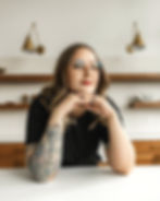 Picture of Sara Wetmore, a white woman with light brown hair and tattoos on her forearm, wearing a black shirt and glasses with her head resting on her hands