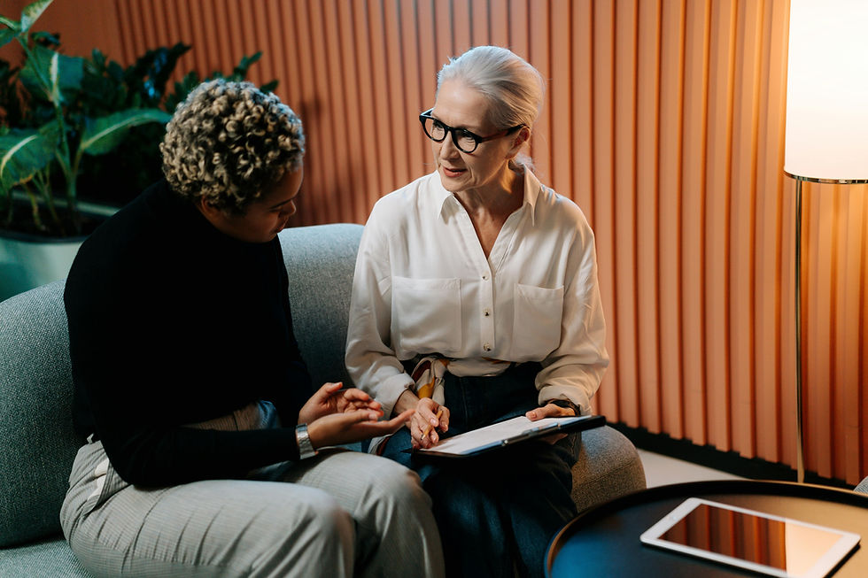 An woman with white hair and glasses engages in a conversation with a younger woman while holding a clipboard. They sit together on a couch in a warmly lit, modern office setting, suggesting a mentoring or coaching session. Representing the perspective of Matthew Schissler on transparent communication in leadership.