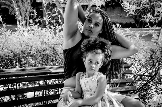 Retrato em preto e branco, mãe e filha sentadas em um banco, a filha ao colo, na sombra de uma árvore no parque da cidade.