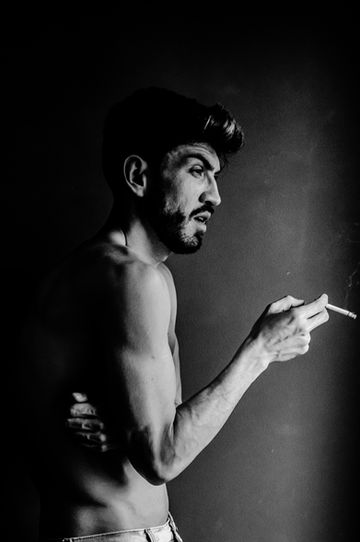 Retrato em preto e branco, meio corpo, modelo masculino jovem com porte atlético, de perfil, olhar pensativo com um tabaco na mão