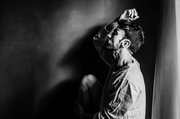 Retrato em preto e branco, meio corpo, modelo masculino jovem com porte atlético, de perfil, sentado com o joelho erguido, passando a mão no cabelo.