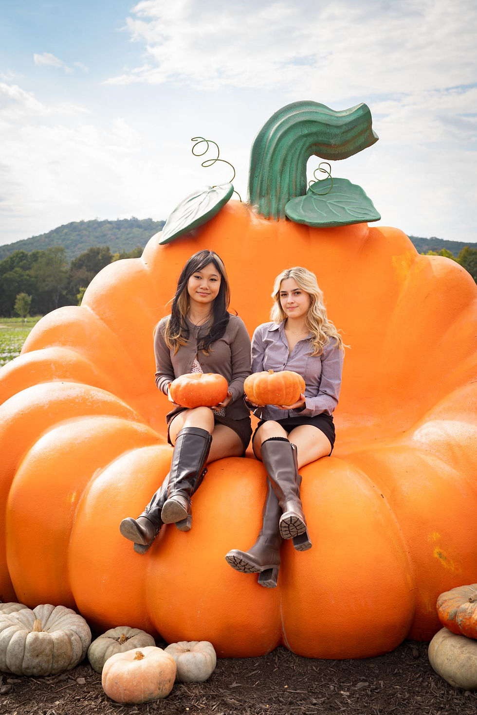 Two friends hold pumpkins as they sit on a giant pumpkin shaped like a bench.