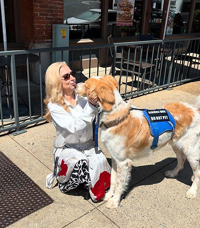 Crystal McDaniel, founder of ADA Paws Pass by Crystal, kneels on a sunny sidewalk in front of a café, sharing a sweet moment with her 105-pound service dog, Big. Crystal rocks a bold red-and-black floral skirt, crisp white blouse, and oversized sunglasses—glamour meets grit. Big, decked out in his bright blue “Service Dog – Do Not Pet” vest, lovingly leans in for a kiss, proving once again he's not just a good boy—he’s the best boy.