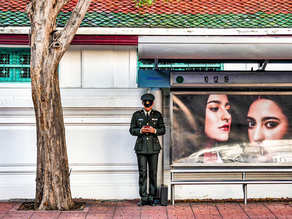A Thai soldier in a formal green uniform stands at a bus stop, looking down at his smartphone, juxtaposed with a large advertisement featuring the faces of two women.