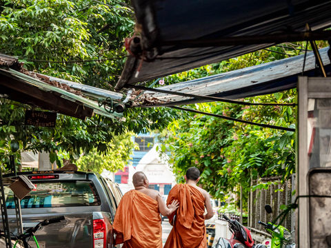 Two Buddhist monks in orange robes walking down a narrow, shaded alleyway in Bangkok, passing parked motorcycles and a pickup truck.