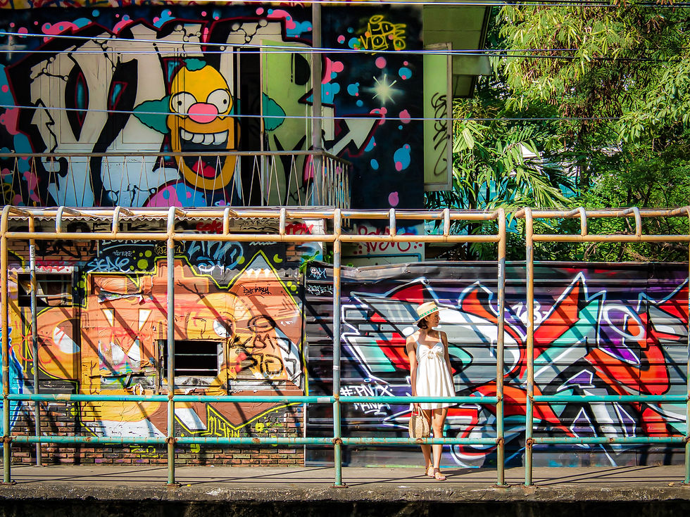 A woman in a white dress and sun hat stands on an urban walkway in front of a massive wall covered in vibrant, multi-layered graffiti and street art.