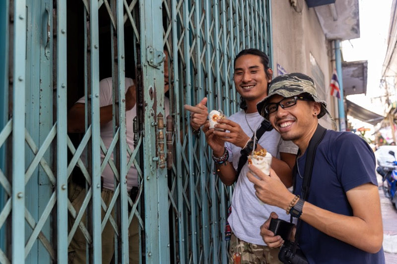 Two travellers enjoying Thai street snacks with their photographer friend in Bangkok