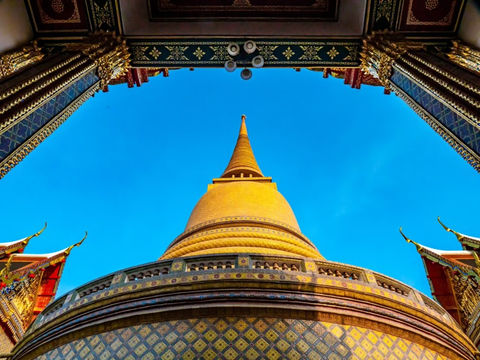 A low-angle view looking up through an ornate Thai temple archway, which frames a large, brilliant golden stupa (chedi) against a clear, deep blue sky.
