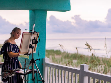 Woman painting at an easel on a beachside porch. She wears a striped dress, surrounded by a teal pillar and ocean view at sunset.