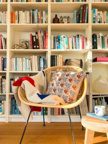 Cushion with pine bloom print in wicker chair with knit blanket and bookcase in the background