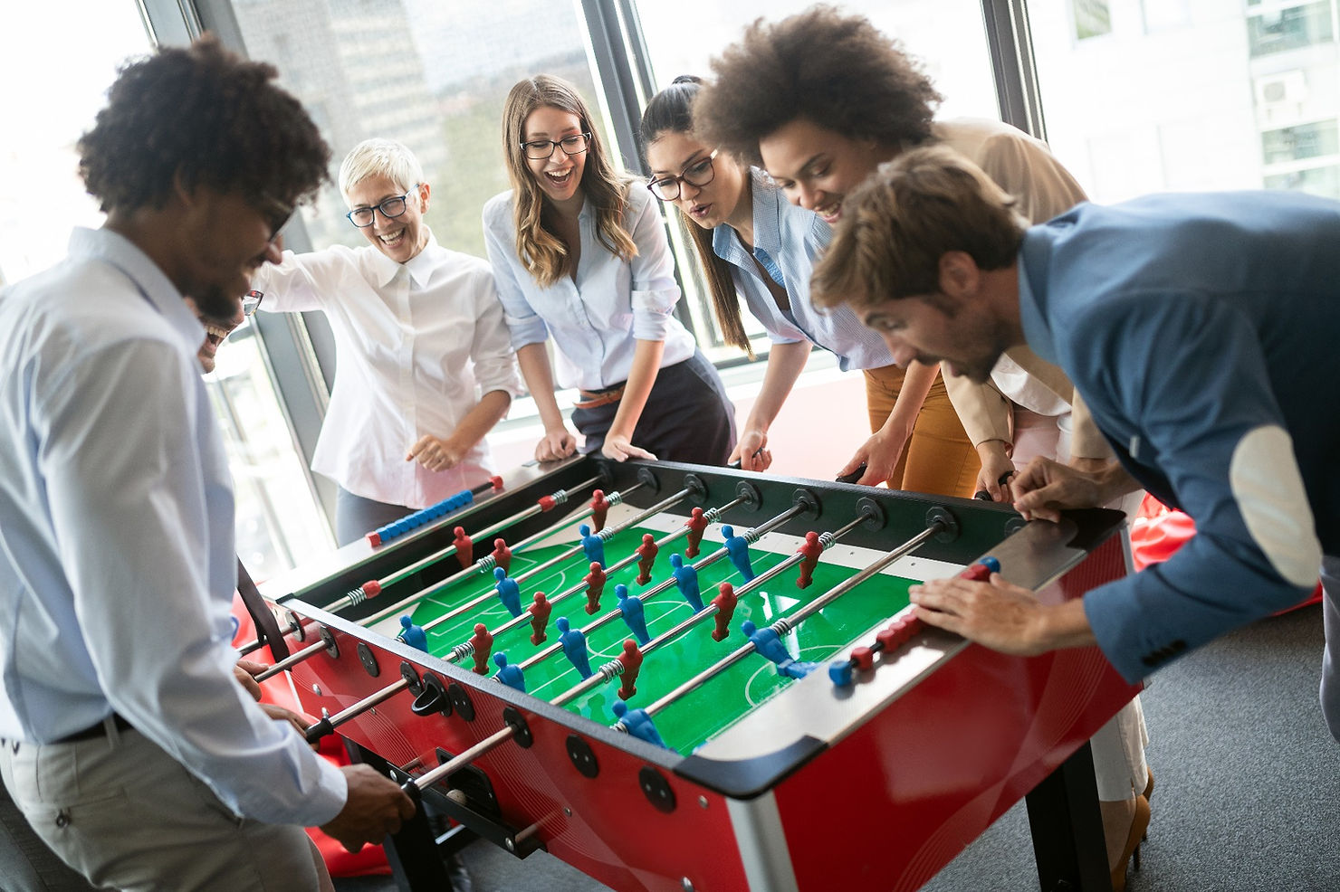 People enjoying a foosball game in a bright office space. They're smiling and focused, with a green field and red foosball table.