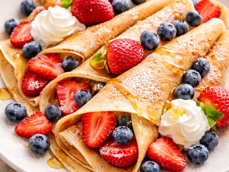 Crepes topped with strawberries, blueberries, and whipped cream on a white plate. Honey drizzles and powdered sugar add sweetness.