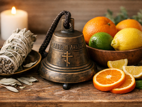 Bronze bell with Cyrillic text and cross sits on a wooden table with sliced citrus, sage bundle on a dish, and lit candle in background.