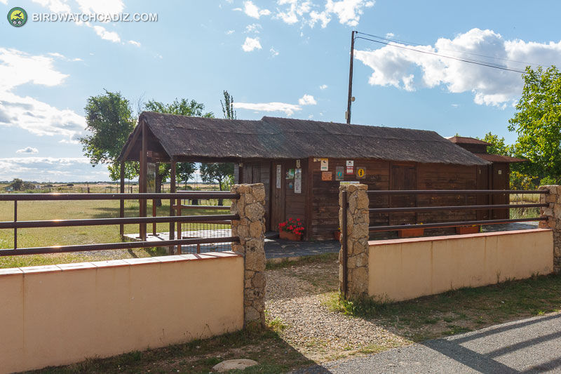 Rustic thatched wooden cabin behind a low wall and gate under a bright blue sky.