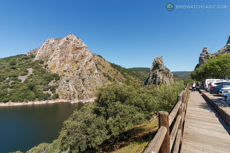 Rocky cliffs and a calm river with green foliage. A wooden walkway to the right has parked cars and a person walking under a clear blue sky.