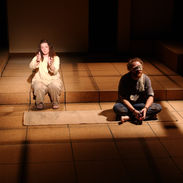 A captive man sits on the floor while a woman talks from a different space