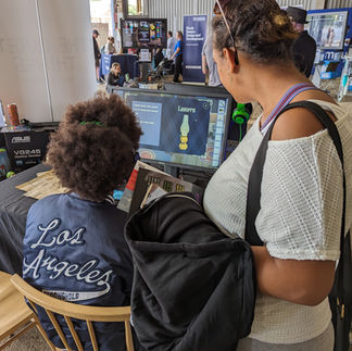 A person sits at a table playing Violet's Vision while a parent watches over them. They play on a PC. They wear black headphones over curly black hair.