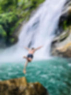 Man jumping off rocky ledge into turquoise pool below a waterfall, surrounded by lush greenery. Joyful, adventurous mood.