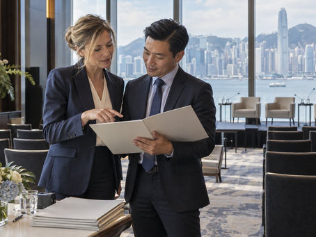 An overseas finance speaker and Hong Kong event coordinator reviewing a plain programme folder beside Victoria Harbour.