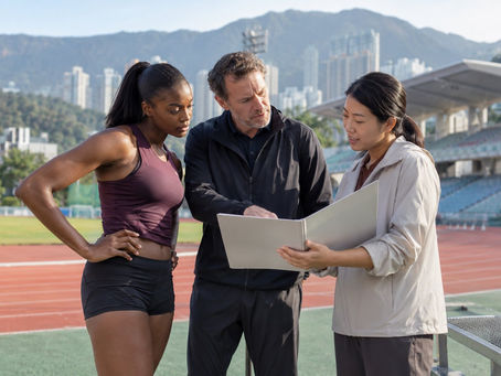 An overseas athlete and coach reviewing an event folder with a Hong Kong coordinator beside an athletics track.