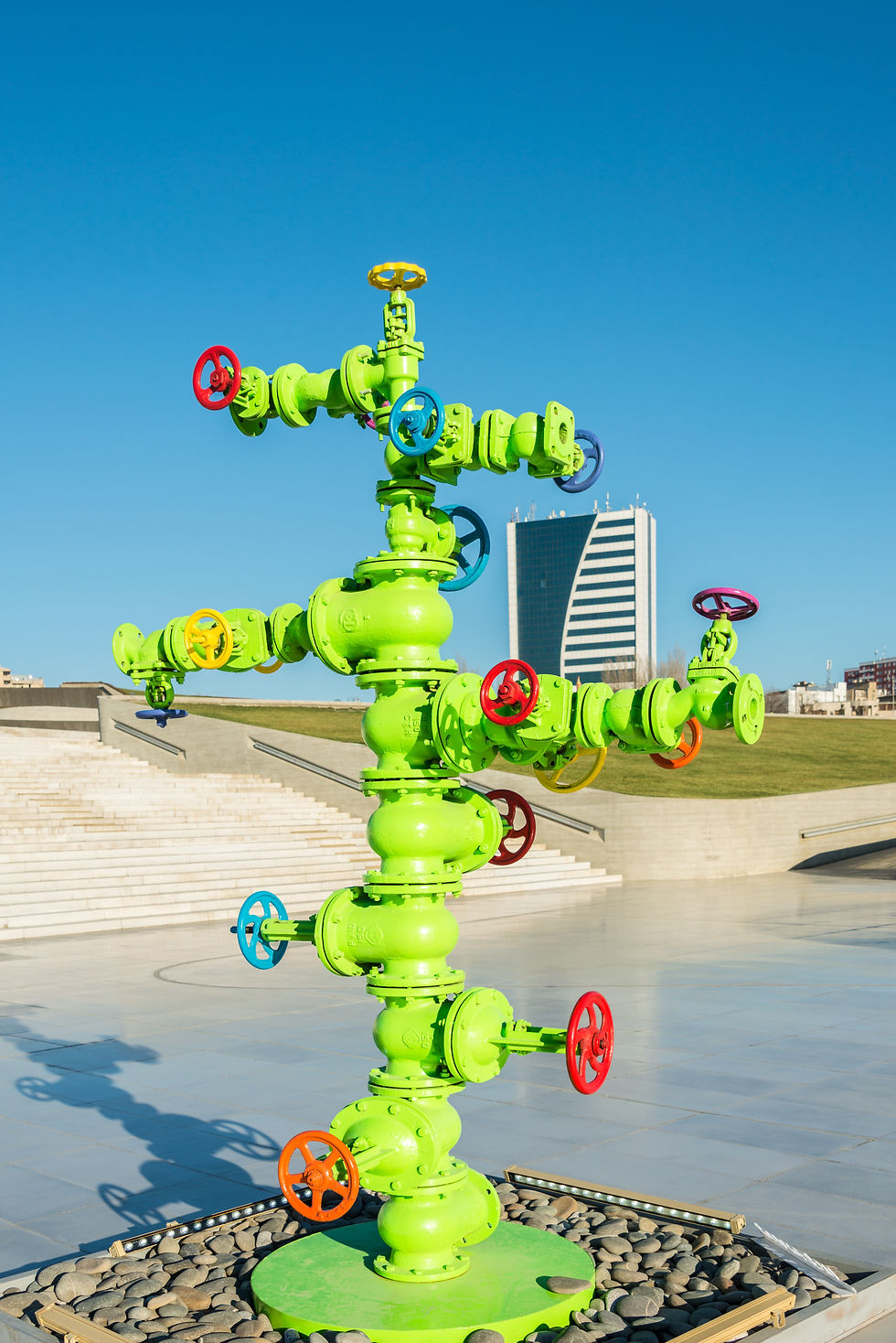 A brightly colored artistic installation of a petroleum Christmas tree painted in vibrant green with colorful red, yellow, and blue valve handles. The structure is set in a modern urban outdoor environment, with a building and landscaped area in the background under a clear blue sky.