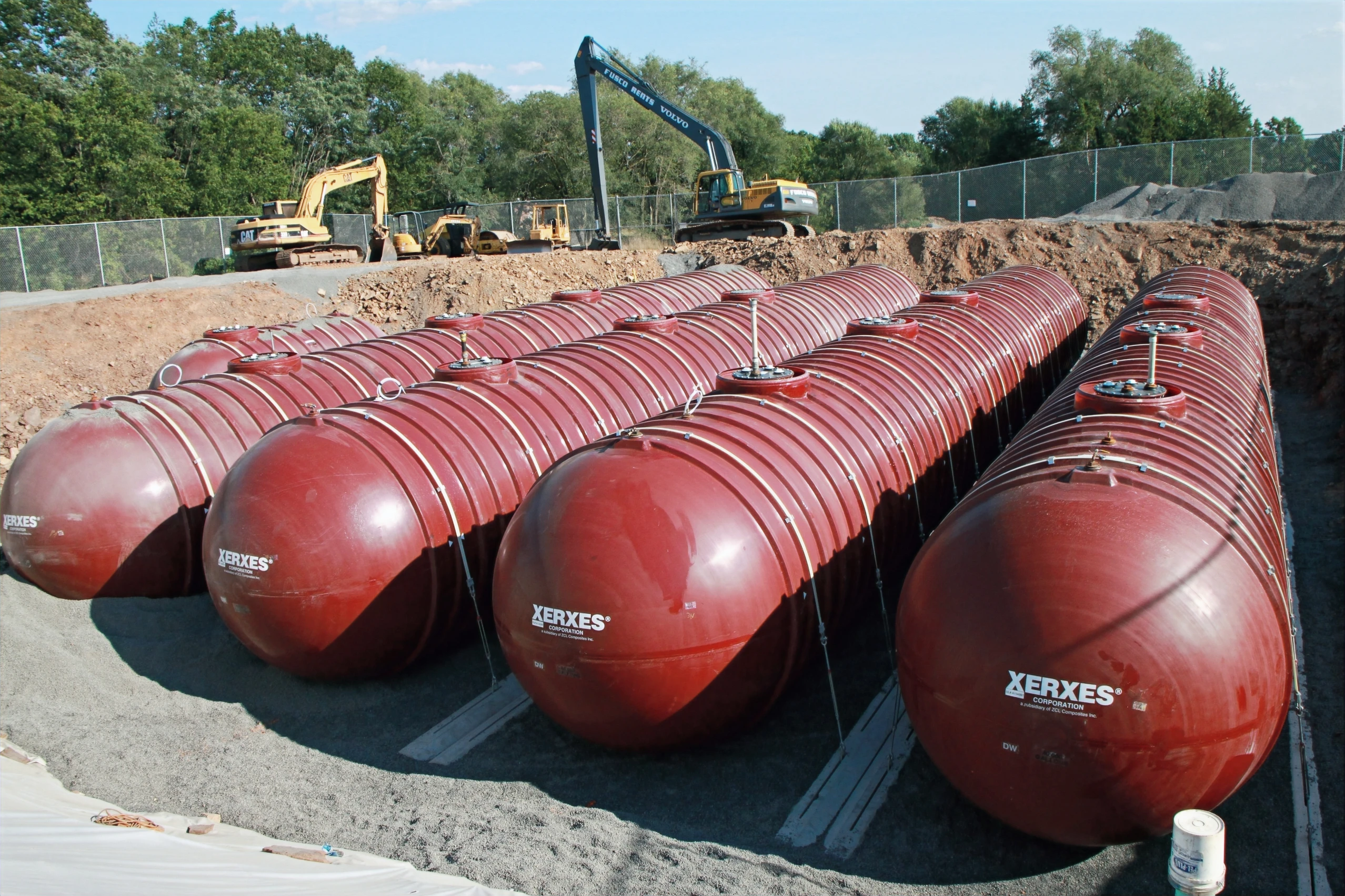 A construction site showing several large, red cylindrical underground storage Xerxes tanks lined up in a trench, with excavators working in the background under a clear blue sky.