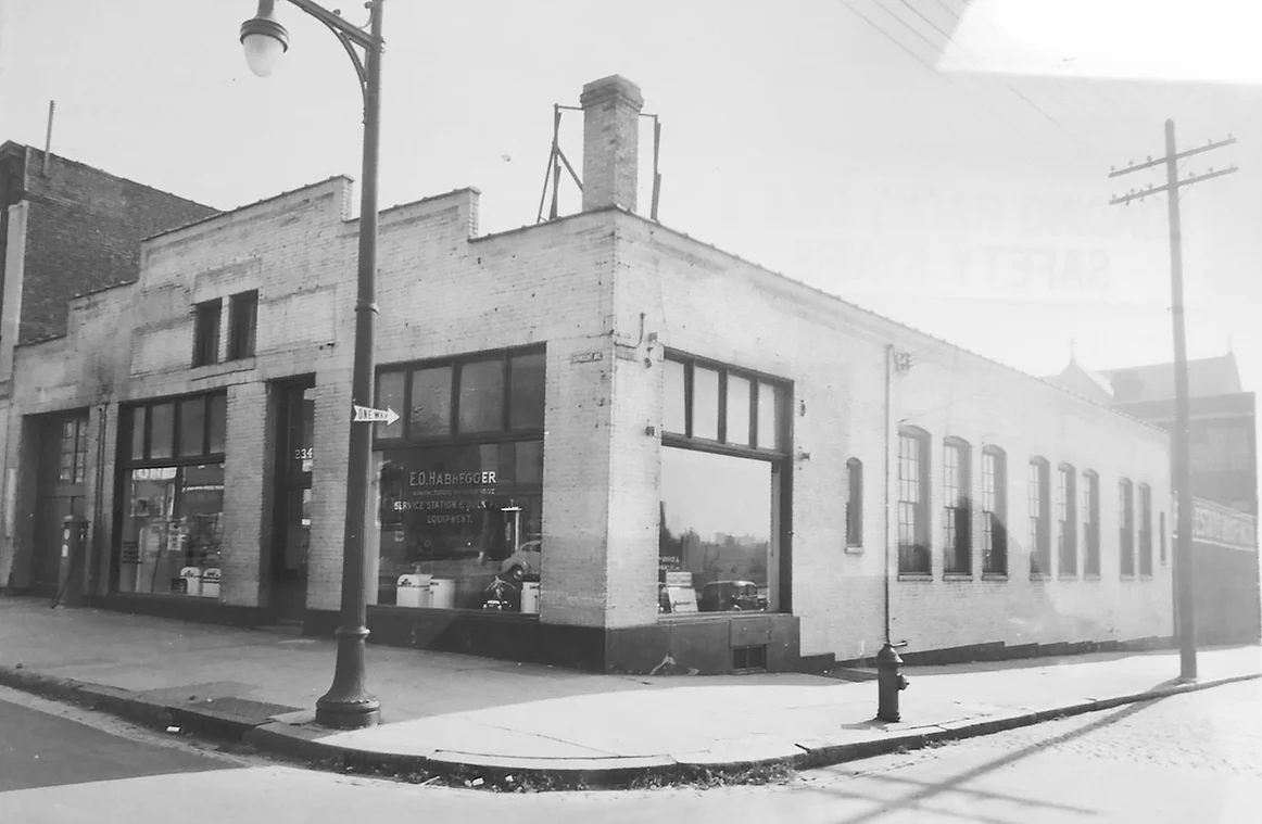 A wide-angle black-and-white photo of a corner brick building with large glass display windows and an "E.O. Habhegger" sign.