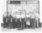 A group of thirteen men in 1940s work attire standing in front of a storefront window featuring a "United We Stand" patriotic banner.