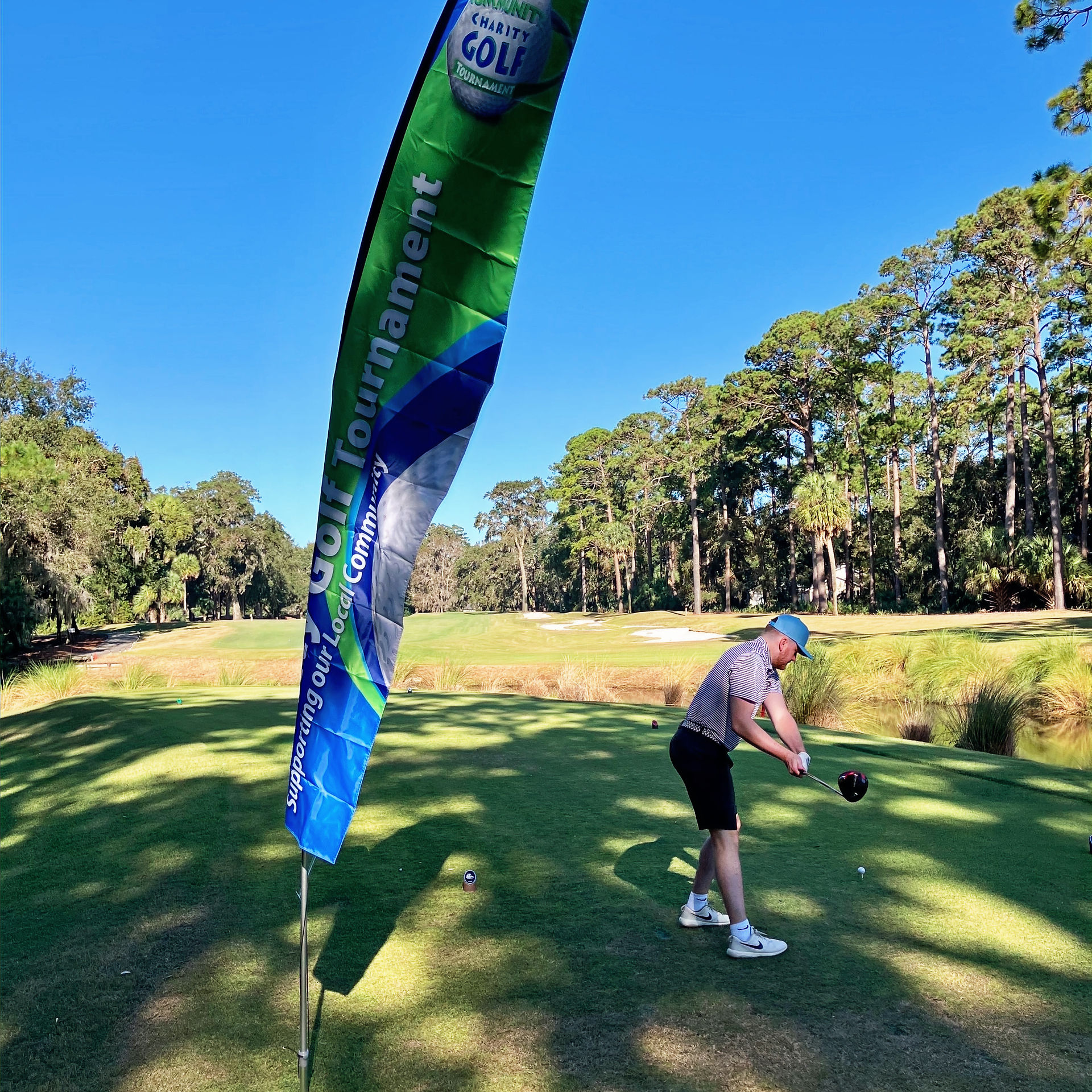 Golfer preparing to swing at Parker's Fueling Community Charity Golf Tournament, with a large tournament banner in the foreground and trees lining the course in the background.