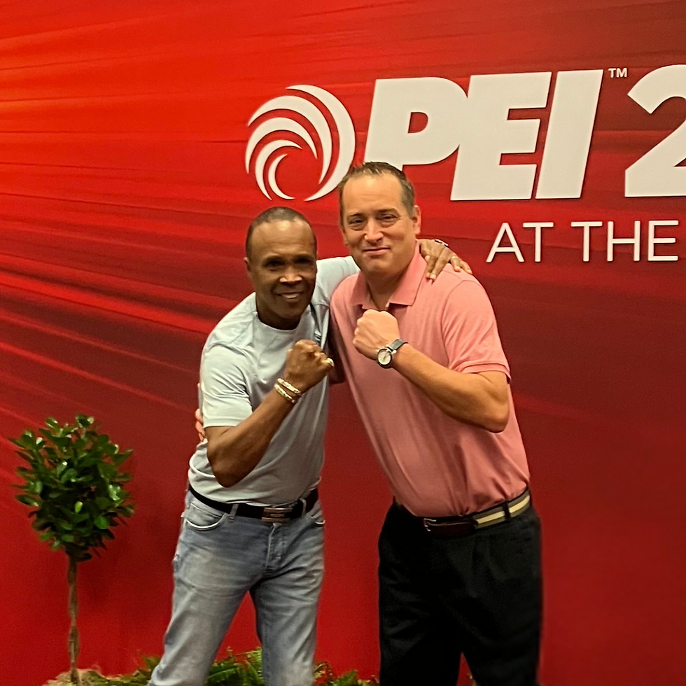 Chris Hagman and Sugar Ray Leonard pose together at the NACS 2022 Trade Show. Both are smiling and making a fist, with a red event backdrop displaying the "PEI" logo behind them.