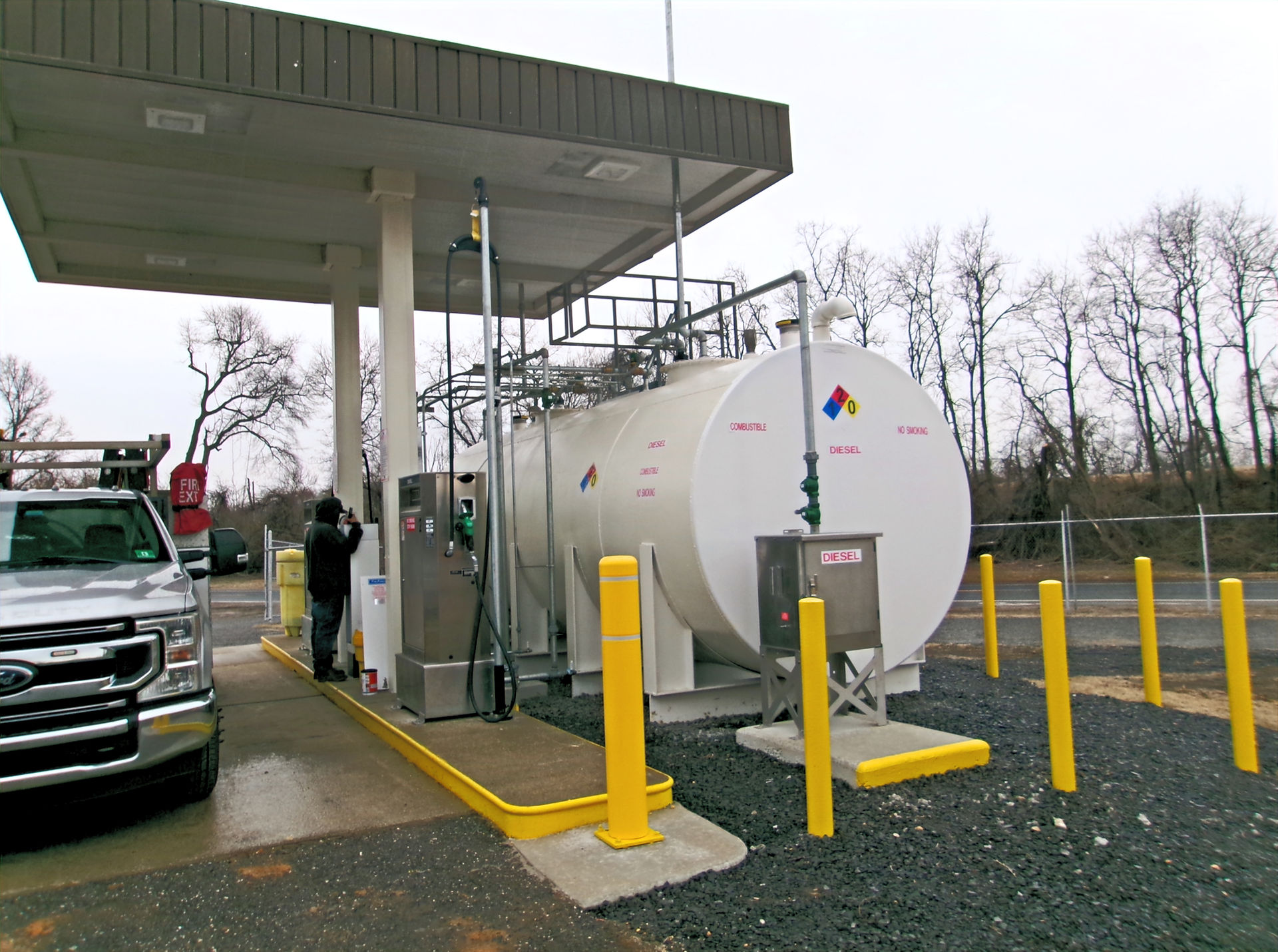 Side view of a newly installed dual aboveground fuel storage tank system under a modern fueling canopy, with bright yellow safety bollards, clean pavement, and updated pump equipment.