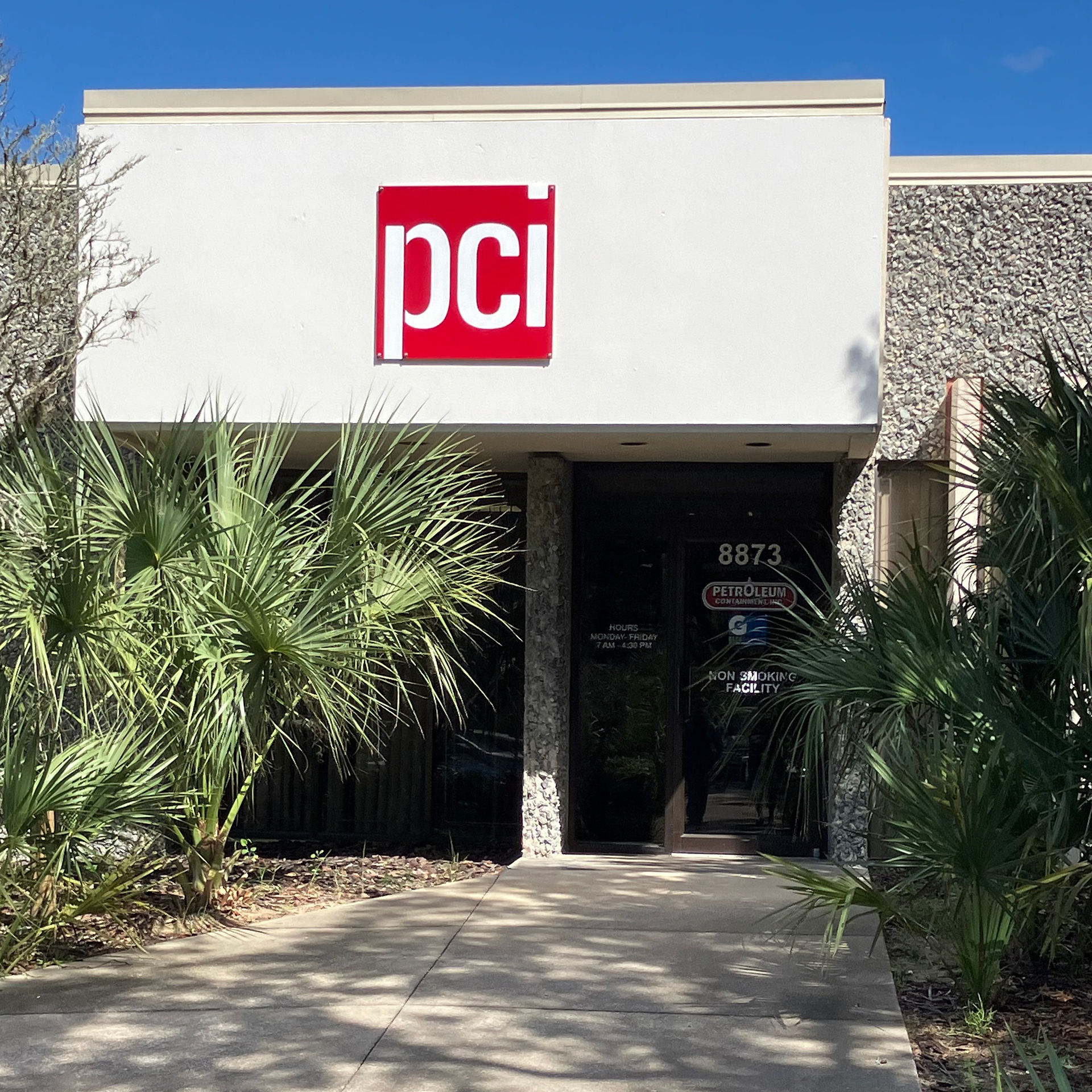Entrance to PCI manufacturing facility featuring the PCI logo prominently displayed above the door, with palm trees and landscaping surrounding the building.