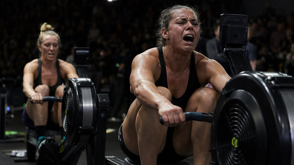 Two female athletes pushing through the final strokes of a high-intensity indoor rowing race, showing visible fatigue and effort.