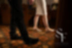A couple walks hand in hand in a church with colorful tiled floors. The woman wears a white dress and heels, the man wears black shoes. Text: Stories Forever Weddings.
