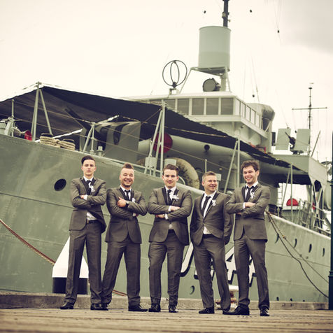 Five groomsmen in suits posing in front of a large ship.
