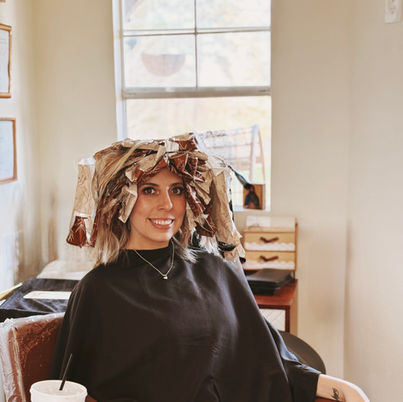 Smiling woman with foils sitting in salon chair holding a drink.