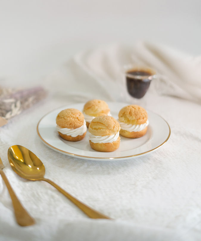 Fotografia de sobremesa francesa, uma massa choux, recheada com creme leve e branco, acompanhada com uma chácara de café em um fundo minimalista branco.