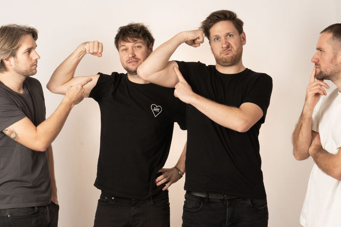 Die Bandmitglieder von „The Flying Fivers“ posieren humorvoll: Zwei zeigen auf die Bizeps-Pose eines Kollegen, während ein anderer nachdenklich schaut.