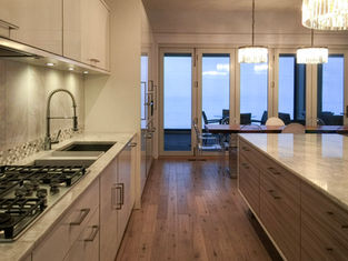 Modern kitchen with beige cabinets, marble countertops, and stainless steel appliances. Bright pendant lights illuminate. Glass doors lead outside.