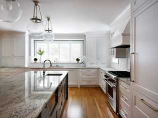 Modern kitchen with white cabinets, wooden floor, and granite countertops. Large window, hanging glass lights, and silver appliances enhance brightness. featuring subzero and wolf, designed by Lifestyle Kitchen Studio