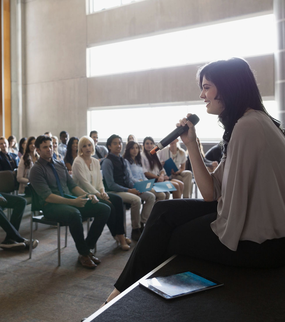 A woman stands on a stage speaking into a microphone to an attentive audience in a bright, well-lit conference room. The scene highlights community learning and sharing knowledge around sustainability. This image supports The Bare Alternative’s Plastic Free July message about staying informed, learning more about low waste living, and engaging with others to create lasting environmental change.